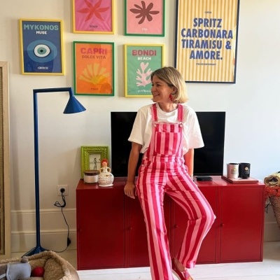 Macacão rosa e branco às riscas com botões metálicos usado por mulher sentada em móvel vermelho com posters coloridos ao fundo