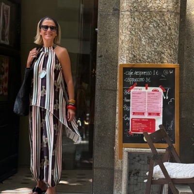 Mulher com macacão riscado com padrão de torneiras e flores, pulseiras e óculos posando junto a uma parede de mármore e menu de café.