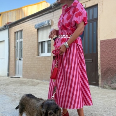 Mulher de camisa rosa com corações vermelhos e saia rosa às riscas, com trela vermelha e cão castanho
