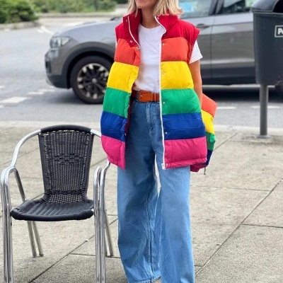 Mulher com colete arco-íris, t-shirt branca, jeans largos, boné azul e óculos escuros na rua junto a cadeira preta e carro.