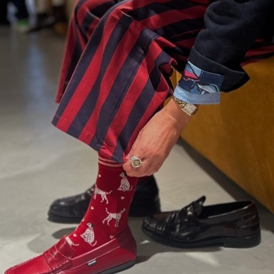 Mãos e pernas de uma pessoa vestindo meias vermelhas com padrão de cães, sapatos mocassins vermelhos e calças com riscas vermelhas e pretas sentado num sofá amarelo.