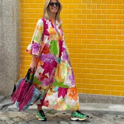 Vestido longo colorido com padrão floral, ténis verde e bolsa com franjas, parede amarela de azulejos no fundo