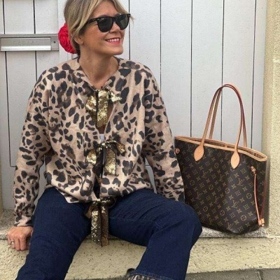 Mulher com camisola de malha padrão leopardo, calças azuis, óculos escuros, lenço vermelho no cabelo e bolsa castanha com padrão monograma.