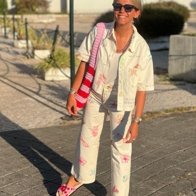 Mulher com conjunto branco estampado, sandálias rosas e mala de croché colorida.