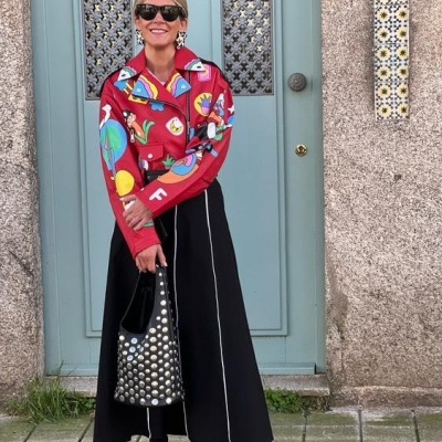 Mulher com casaco vermelho colorido, saia preta, mala e sapatos pretos com bolinhas, em frente a porta azul