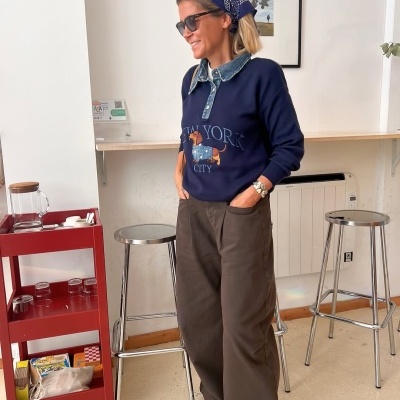 Mulher de camisola azul com estampa de cão e calças castanhas num interior com bancos metálicos e carrinho vermelho.
