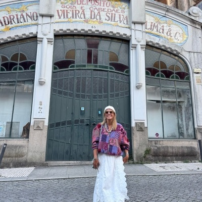 Mulher com vestido branco longo e blusa rosa e lilás com padrão geométrico em frente a edifício antigo com portas de vidro cinza