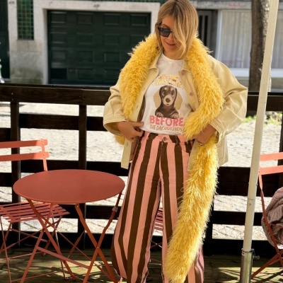 Pessoa com casaco bege e gola felpuda amarela, t-shirt branca com cão e calças listradas rosa e castanho, ao lado de mesa e cadeira vermelha.