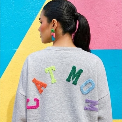 Pessoa de costas com sweatshirt cinzenta e letras coloridas CATMOM e brincos coloridos, perto de parede colorida