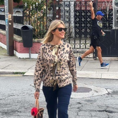 Mulher a caminhar em rua, com casaco de padrão leopardo, calças azuis e sapatos vermelhos, segurando mala castanha com detalhe vermelho.