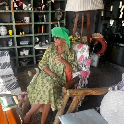 Mulher sentada em cadeira de madeira com vestido verde floral e chapéu verde-limão num espaço com estantes e luminária alta