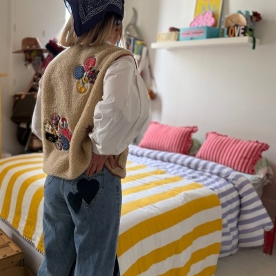 Pessoa com colete peludo bege com apliques coloridos, camisa branca, jeans com corações pretos aplicados e lenço azul branco na cabeça em quarto com cama listrada amarela e branca.