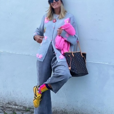 Mulher com roupa cinzenta e rosa, com sapatilhas amarelas e mala castanha em parede de fundo