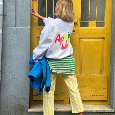 Pessoa de costas com camisola cinzenta, calças às riscas amarelas, ténis azuis Nike, segurando casaco azul, em frente a porta amarela.