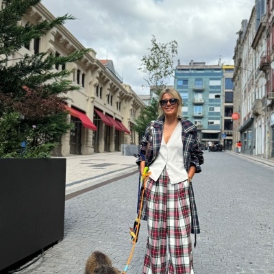 Mulher com roupa xadrez e colete branco a passear cão numa rua urbana