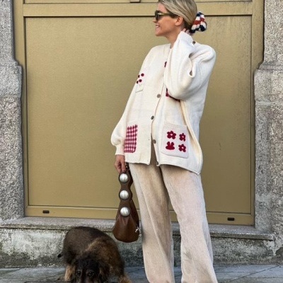 Mulher de cardigã creme com padrões em vinho e rosa, calças creme largas, sapatilhas castanhas, lenço no cabelo e mala castanha, com cão castanho junto.