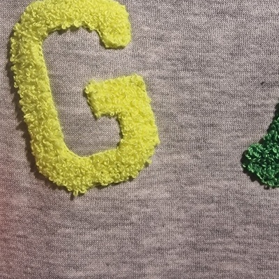 Close-up de tecido cinzento com letra G felpuda amarela e parte de letra verde.