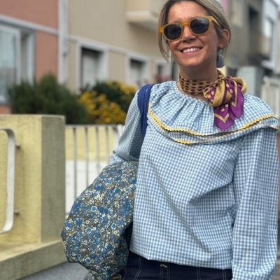 Mulher vestindo camisa xadrez azul e branca com folhos amarelos, cachecol colorido, saia preta, óculos de sol castanhos e bolsa azul floral.