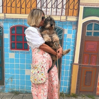 Pessoa com macacão rosa floral segurando cão junto a mural colorido de azulejos e portas