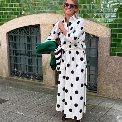 Mulher com vestido branco às bolinhas pretas segurando casaco verde em frente a parede de azulejos verdes