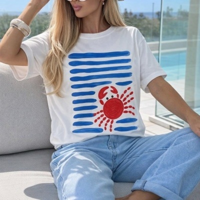 Mulher com t-shirt branca com caranguejo vermelho e linhas azuis, calças de ganga, chapéu de palha e óculos de sol
