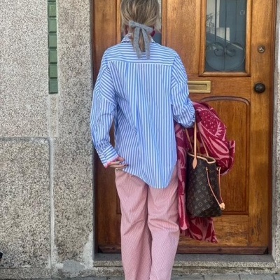 Pessoa de costas com camisa riscada azul e branca e calças cor-de-rosa segurando mala castanha junto a porta de madeira.