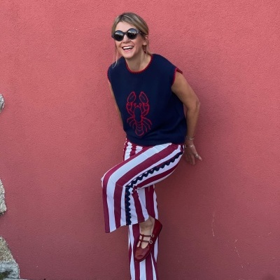 Mulher com camisola de malha azul com lagosta vermelha, calças riscas vermelhas e brancas e sapatos vermelhos junto a parede rosa.