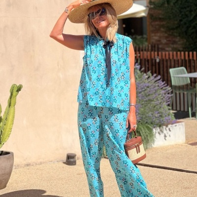 Mulher com conjunto azul turquesa floral, chapéu de palha e óculos de sol, segurando bolsa marrom clara, em ambiente externo com plantas e cadeiras.