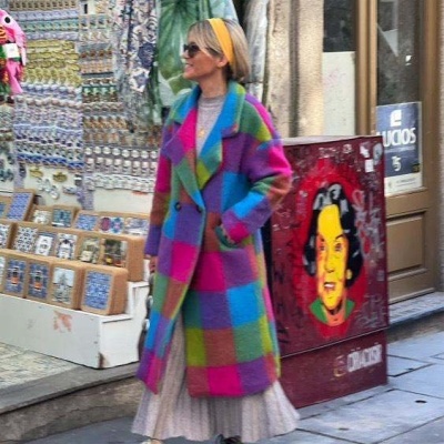 Mulher com casaco xadrez colorido, saia plissada e ténis numa rua com arte e souvenirs.