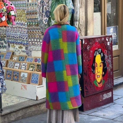 Pessoa com casaco colorido comprido numa rua ao lado de loja com postais expostos