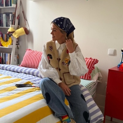 Mulher vestindo colete bege peludo bordado, camisa branca e calças de ganga com padrões sentada numa cama colorida num quarto