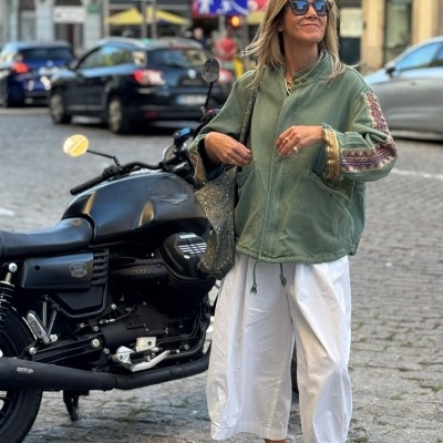 Mulher com casaco verde bordado, calças brancas largas, sapatos bege com atacadores verdes, óculos escuros, bolsa prateada, junto a mota preta numa rua urbana.