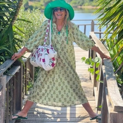 Mulher com vestido verde claro floral, chapéu e sandálias verdes, mala branca floral, em passadiço de madeira com vegetação e lago