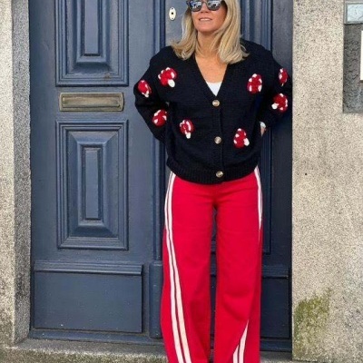 Mulher com cardigan preto de cogumelos vermelhos e calças desportivas vermelhas com riscas brancas, em frente a porta azul escura.