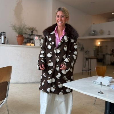 Mulher sorridente com casaco castanho e padrão de ovelhas, vestido branco e camisa rosa, em café moderno e iluminado