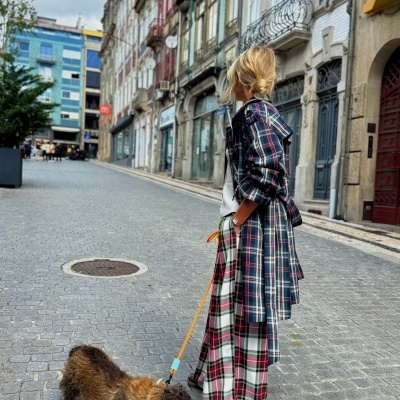 Pessoa com casaco e calças xadrez seguran a trela de cão castanho num espaço urbano