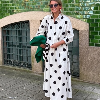 Vestido comprido branco com bolinhas pretas, sapatos pretos e acessórios coloridos em cenário urbano com parede verde.