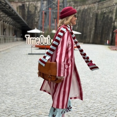 Mulher com casaco às riscas cor-de-rosa e vermelho, boina vermelha, cachecol às riscas, saco castanho, saia azul com flores e mocassins castanhos em calçada.