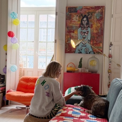 Sala de estar com sofá cinza, cadeira laranja, estante vermelha, quadro colorido e mulher com cão preto