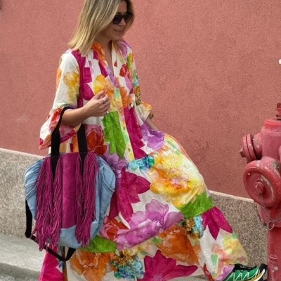 Mulher com vestido longo floral colorido, mochila azul com franjas rosa e tênis verde, sentada junto a parede rosa e hidrante vermelho