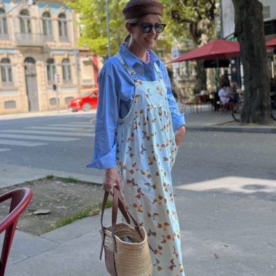 Pessoa em ambiente urbano com vestido comprido branco com padrão de cães, camisa azul clara e bolsa de palha.