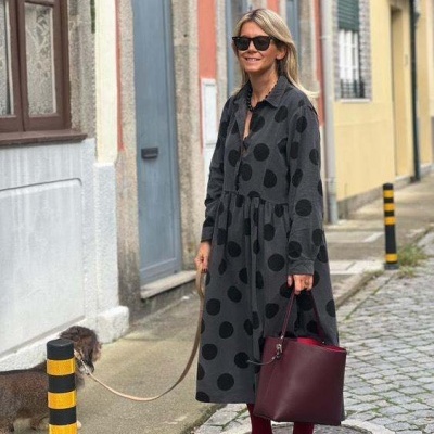 Mulher com vestido preto de bolinhas, óculos escuros e bolsa vinho numa rua com cão