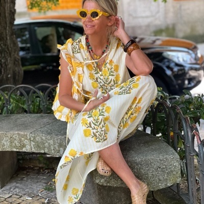 Mulher sentada num banco de pedra com vestido longo floral amarelo, óculos de sol amarelos, colar colorido e alpercatas naturais