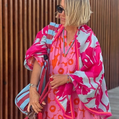 Mulher com vestido florido rosa e laranja, xale rosa, bolsa às riscas, óculos escuros, pulseiras e colar, em frente a parede de ripas de madeira