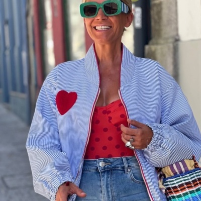 Mulher com casaco riscado azul e branco com coração vermelho, vestido vermelho às bolas, saia de ganga, óculos verdes e mala de palha colorida