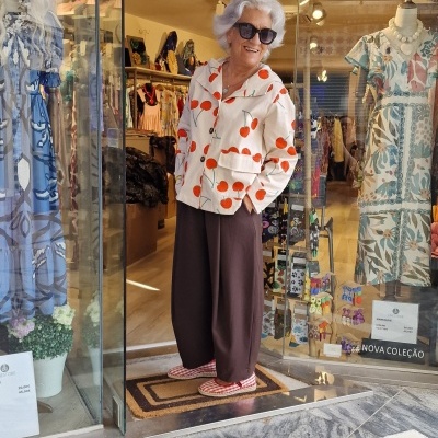 Mulher idosa com blusa branca de cenouras laranja e calças castanhas na entrada de loja de roupas