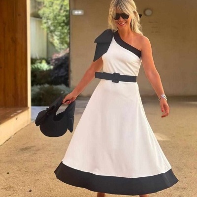 Mulher usa vestido branco com detalhes pretos e segura chapéu preto ao ar livre.