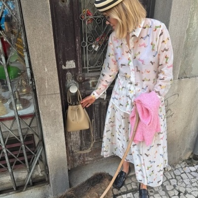 Mulher com vestido branco estampado e chapéu listrado junto a porta antiga com cão.