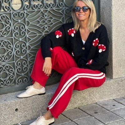 Mulher com cardigan preto com cogumelos vermelhos, calças vermelhas com riscas brancas e sapatos brancos sentada em pedra junto a grade metálica cinza