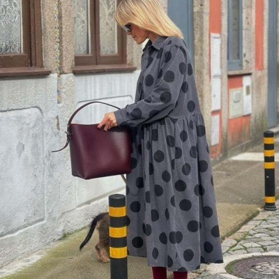 Mulher com vestido cinza de bolinhas negras segurando mala vinho numa rua de calçada e edifícios antigos.